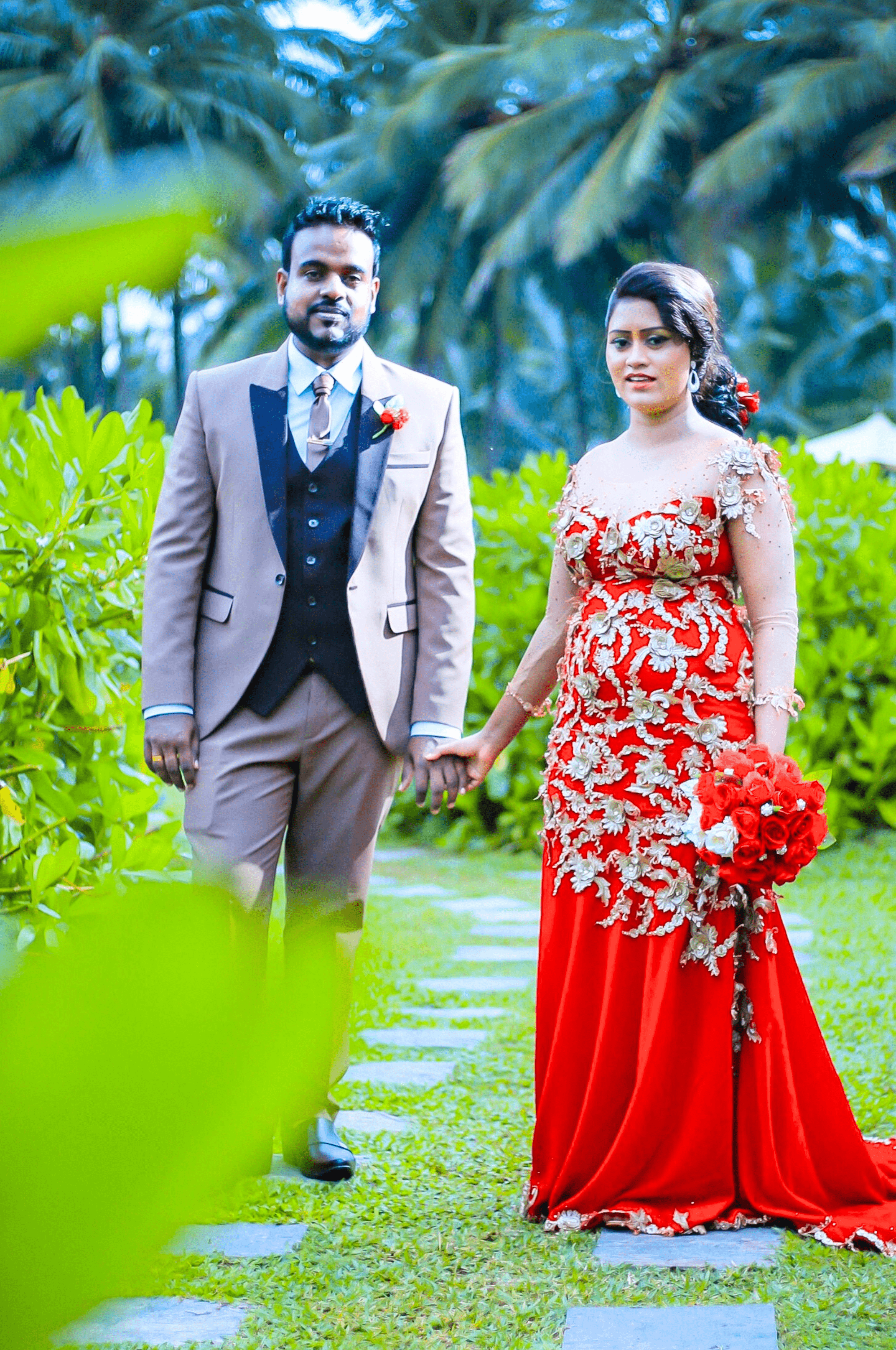 Homecoming wedding couple holding hands in lush greenery background, bride wearing a red wedding frock.